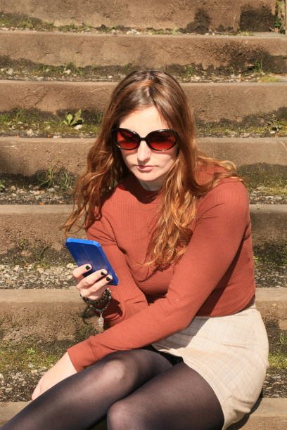 A Caucasian woman texting on a mobile phone while sitting on steps outdoors. She is wearing sunglasses, brown sweater, beige mini skirt, and black stockings.